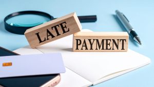 LATE PAYMENT（支払いの遅延）のブロックと虫眼鏡が置かれ、未払い金の支払いがないため督促状の送付や法的措置を検討する様子