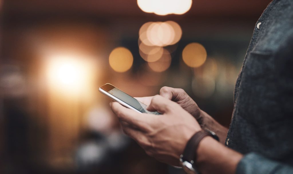ぼやけた背景の中でスマートフォンを操作する男性の手元。美容室の運転資金に関する情報を検索したり融資のオンライン申請を行ったりしている様子