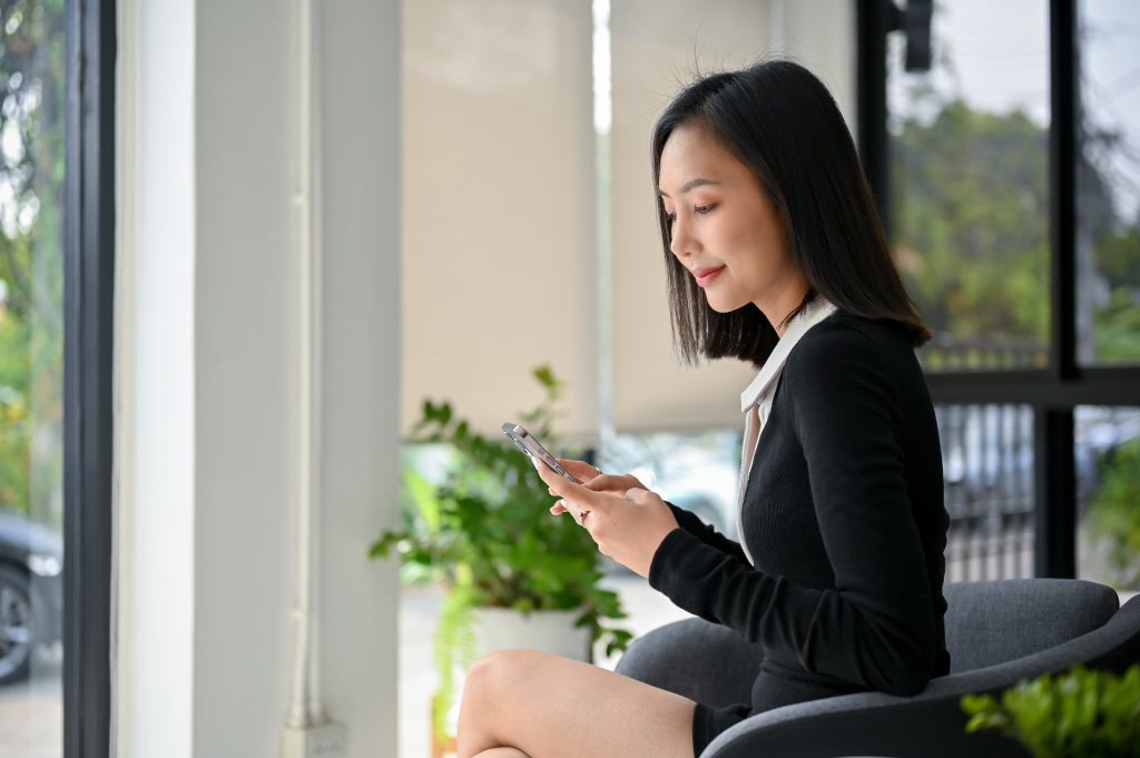 窓際の席でスマートフォンを操作する女性。ファクタリングに変わる選択肢としてAirキャッシュを検討している様子