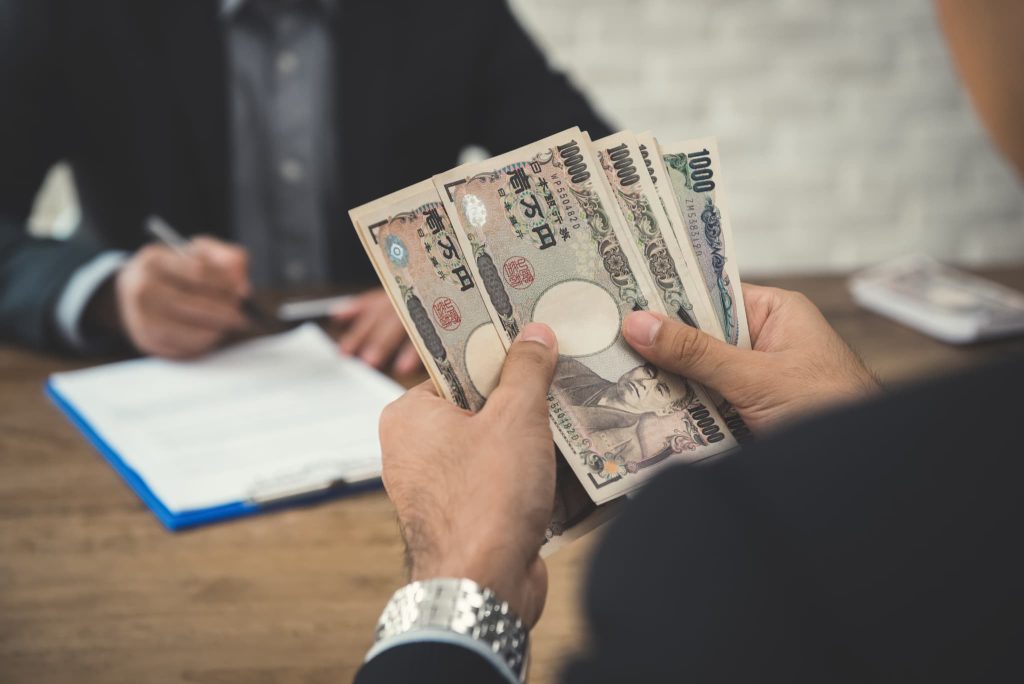 一万円札などの現金を数える手元と、奥で契約書にサインをする人物。即日融資やファクタリングなど、急な支払い対応のために現金を確保する運転資金調達の完了シーン