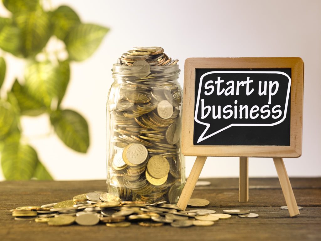 「start up business」と書かれたミニ黒板と、瓶に詰められた大量のコイン。飲食店の開業資金の準備や、創業時の資金調達を象徴するイメージ