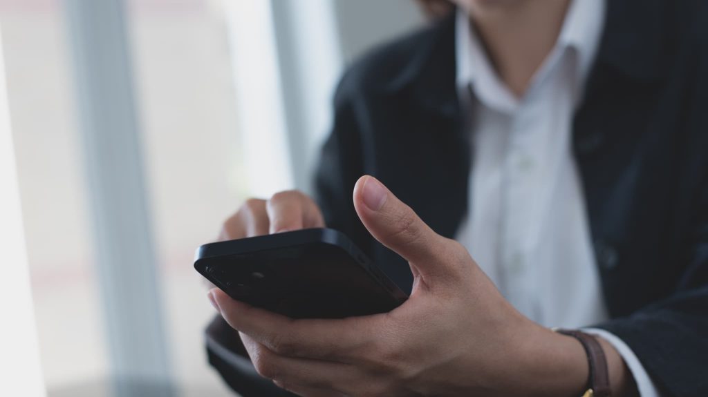 スマートフォンを手に持ち画面を操作しているスーツ姿の人物。債務超過の解決策を調べたり、金融機関からの連絡を確認しているビジネスシーン