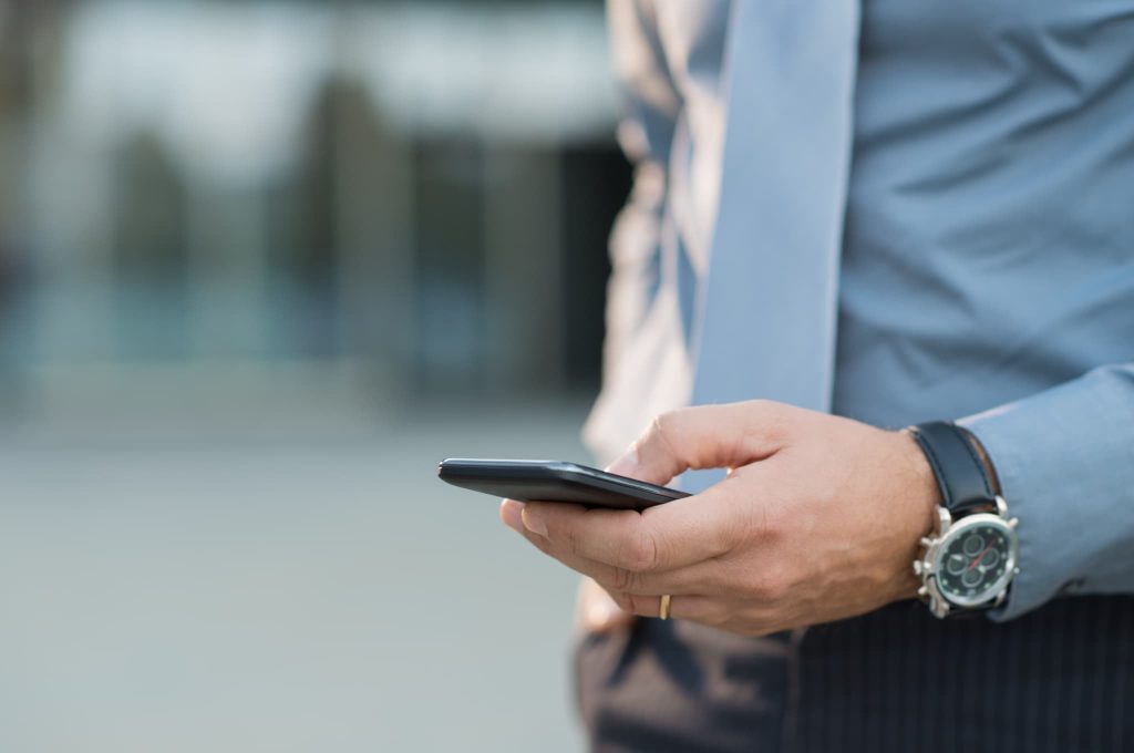 ビジネス街でスマートフォンを操作するスーツ姿の男性。場所を選ばずに申請可能なクラウドファンディングや、返済不要の資金調達に関する最新情報を検索しているビジネスシーン