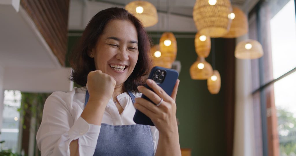 スマートフォンで開業資金の着金を確認し店舗内でガッツポーズをして喜ぶ女性オーナー