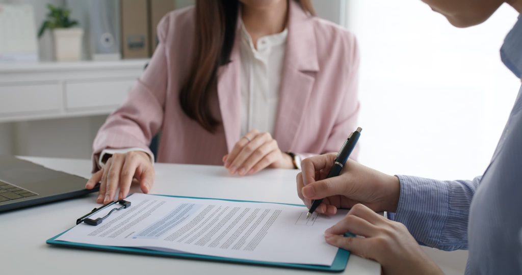オフィスのデスクで書類にペンを走らせる女性と対面の担当者。金融機関へ追加融資の申し込みを行ったり契約手続きを進めているビジネスシーン