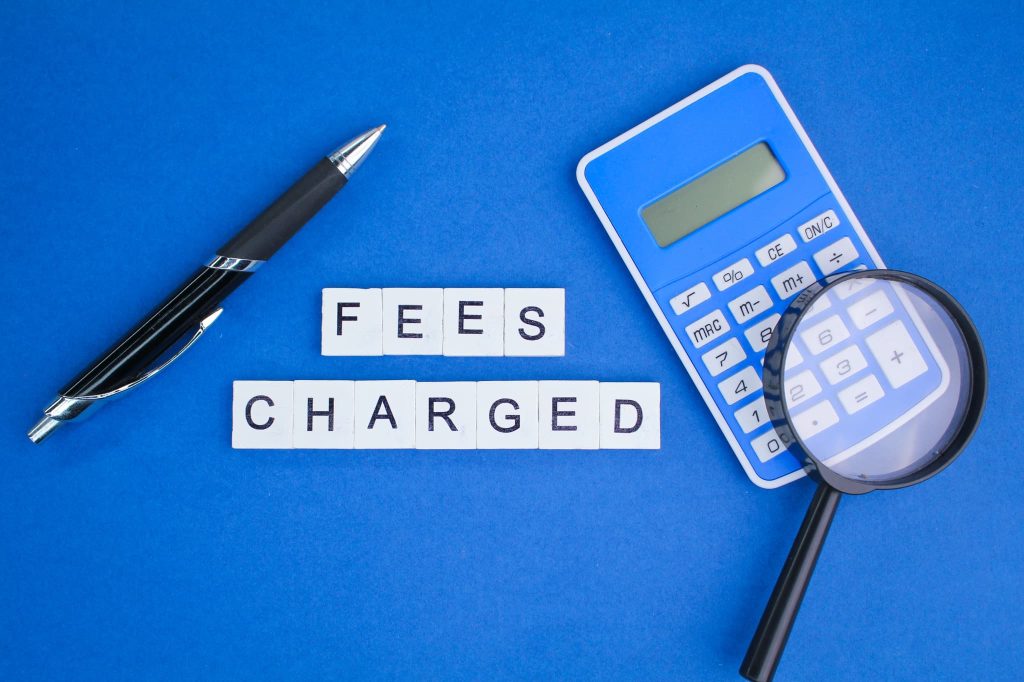 FEES CHARGEDの文字ブロックの横に電卓と虫眼鏡が置かれファクタリングにかかる手数料や費用を詳しく調べているイメージ
