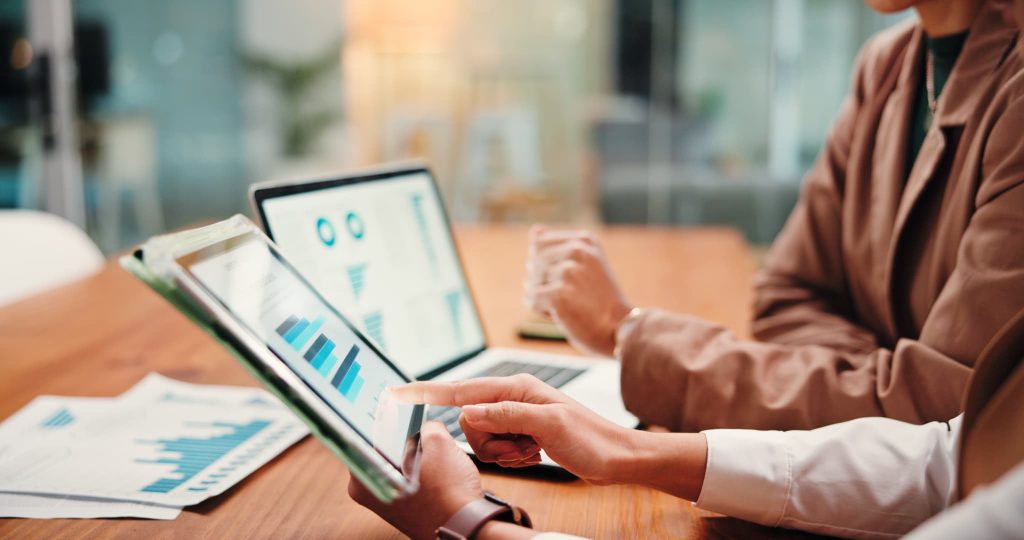 タブレット端末に表示された棒グラフや円グラフを指差して打ち合わせをするビジネスパーソン。将来の現金過不足を予測し資金繰り表を共有するシーン
