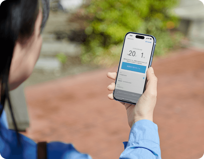 画像：屋外でスマートフォンを眺めている人の様子。スマートフォンには待ち時間と待ち組数、さらに受付ボタンが表示されている。