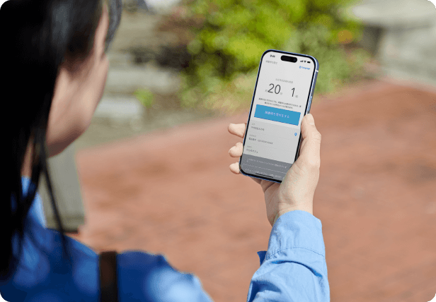 画像：屋外でスマートフォンを眺めている人の様子。スマートフォンには待ち時間と待ち組数、さらに受付ボタンが表示されている。